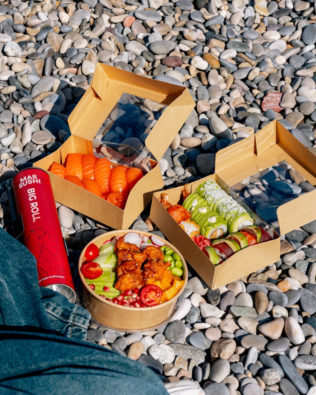 Boîtes Mas Sushi à emporter, poke bowl et Big Roll posés sur les galets de la plage